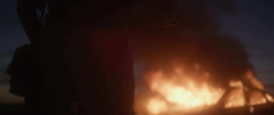 A woman stands next to a burning car at night