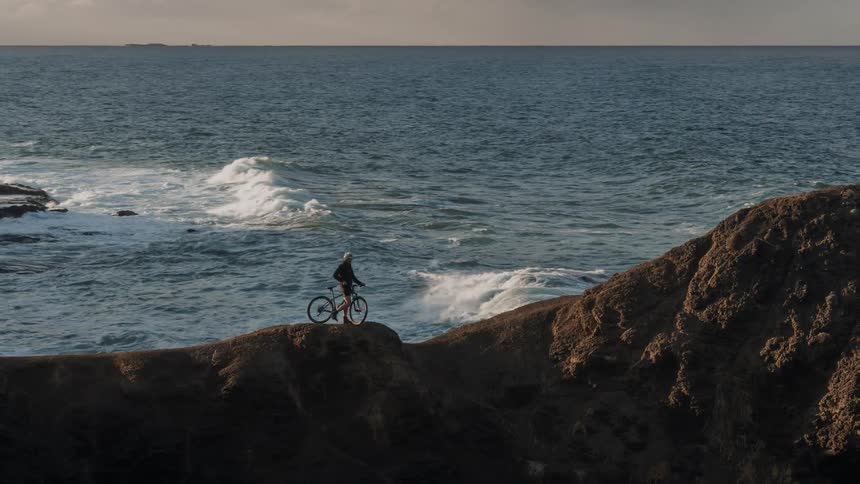 Someone with a big is standing on top of a hill next to the ocean, being filmed from a drone