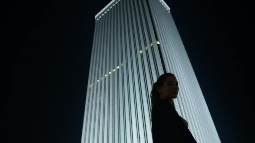 A woman stands in front of a lit skyscraper at night