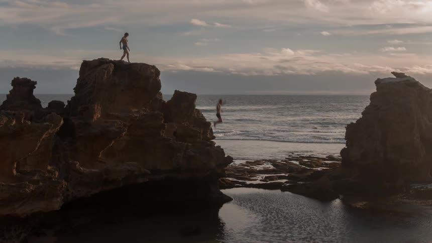 Two men walk to the edge of a cliff , one dives in the water below