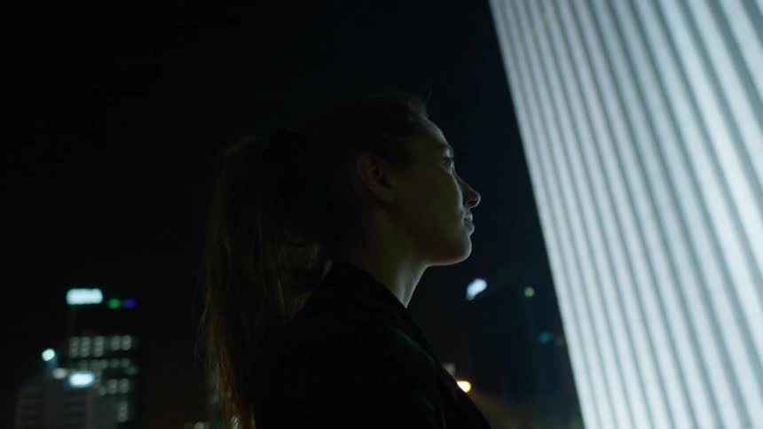 A woman stands in a city at night looking up at a building