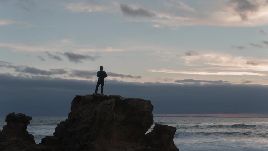 A man stands near a cliff watching the sun set over the ocean