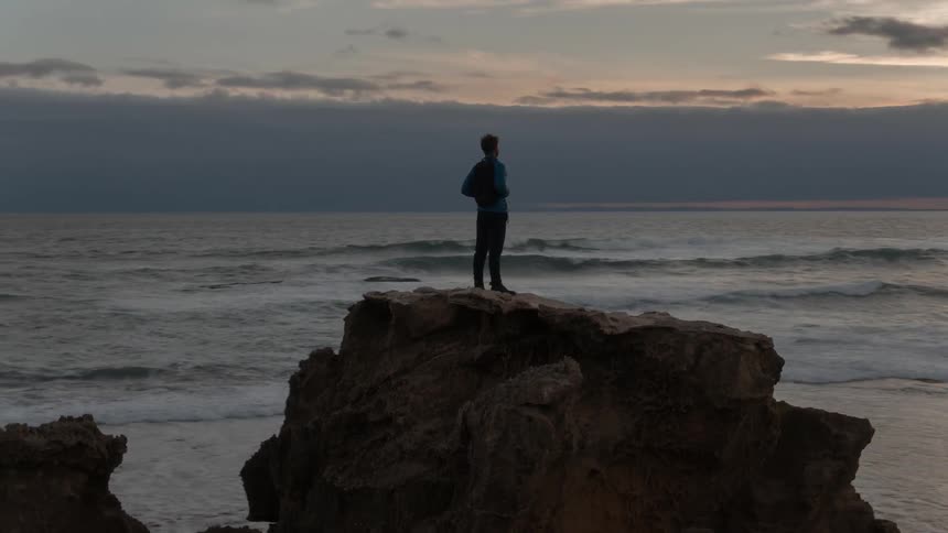 Man stands on a large rock and looks out at the ocean