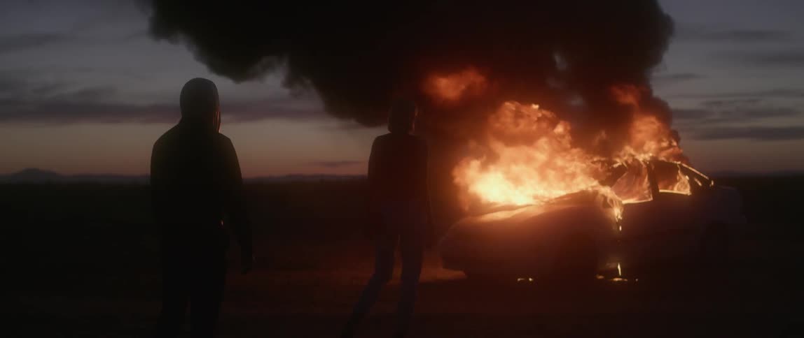 A woman is watching a car burn in the desert and a man walks up behind her