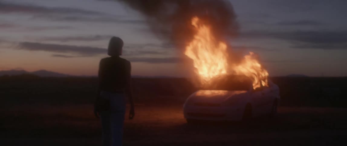 A woman is looking at a car on fire at dusk in the desert