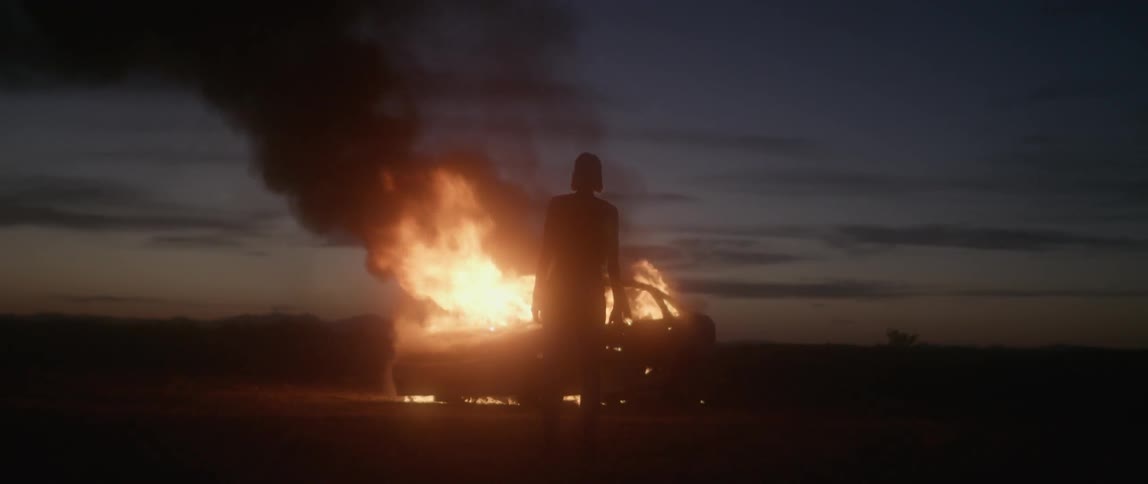 A woman walks over and watches a car burn