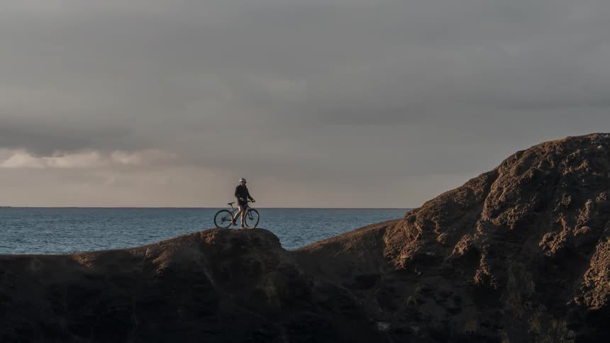 A person with a bike is standing on top of a hill looking at the ocean
