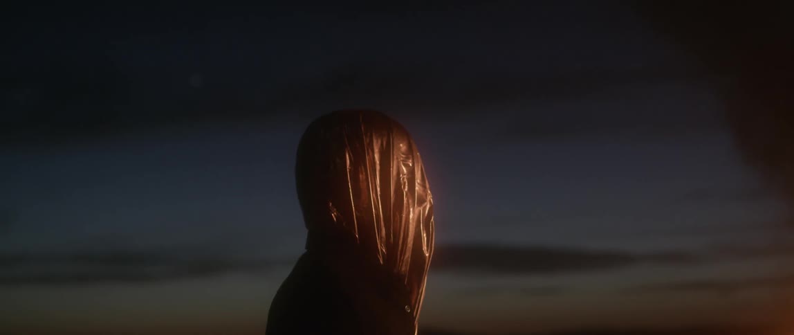 A person wears a bag over their head outside at dusk