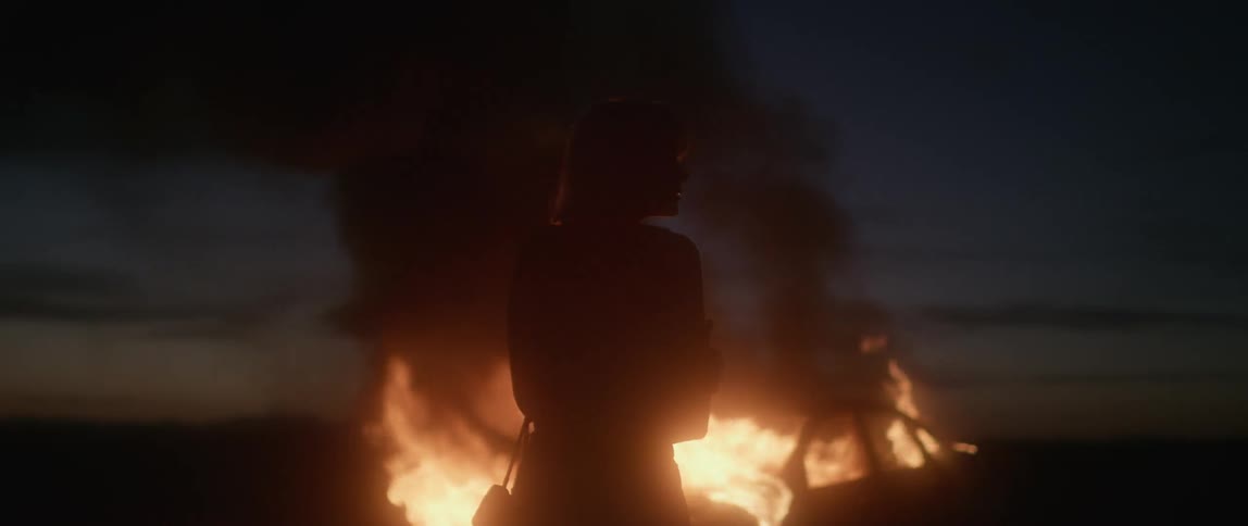 A woman stands in front of a car that is completely aflame