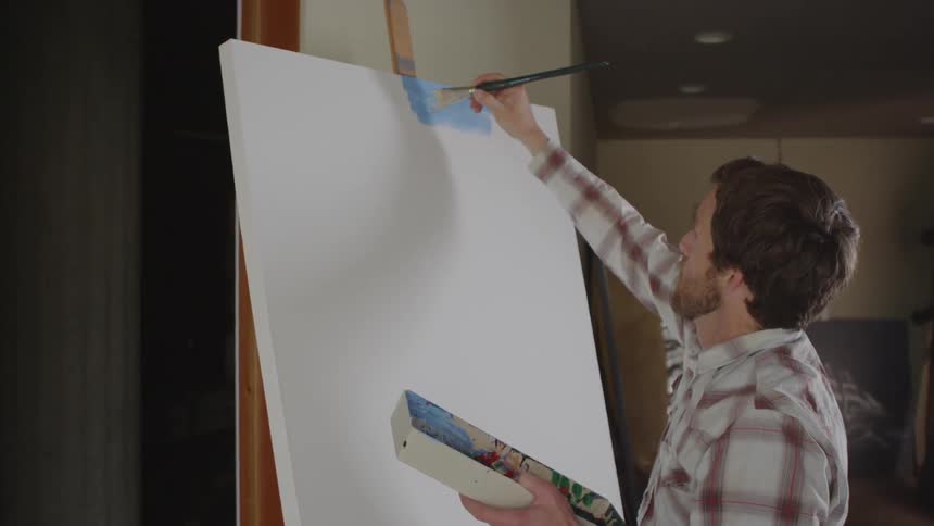A white man stands at an easel and puts blue paint on a large canvas