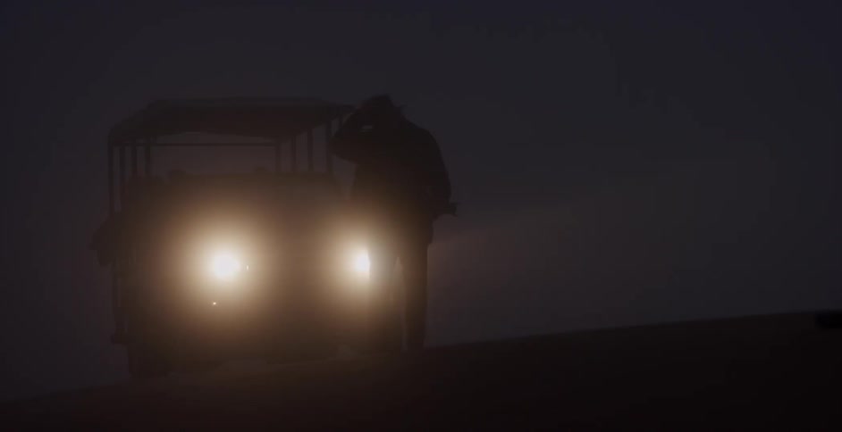 A man walks outside in front of a vehicle with headlights turned on