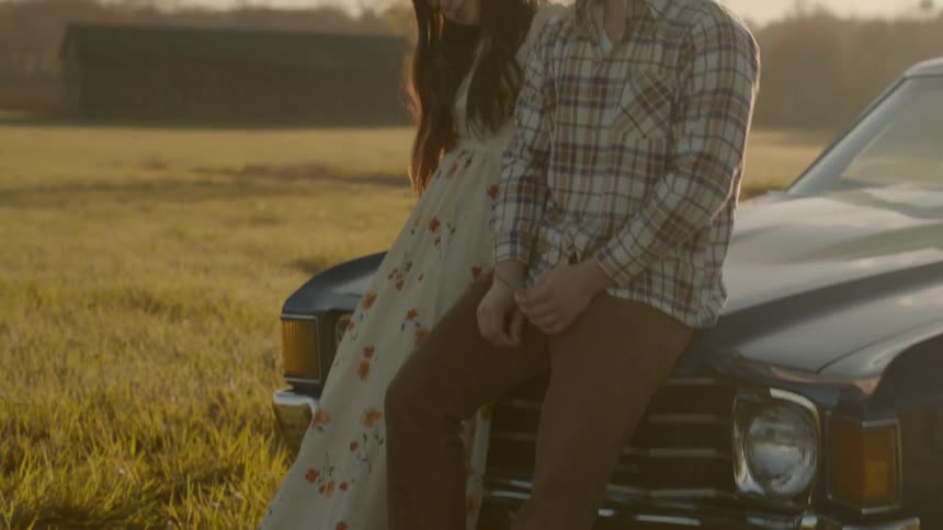 A couple sit on the hood of an old car talking