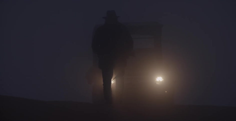 A man in a cowboy hat is walking in front of his car headlights at night while foggy