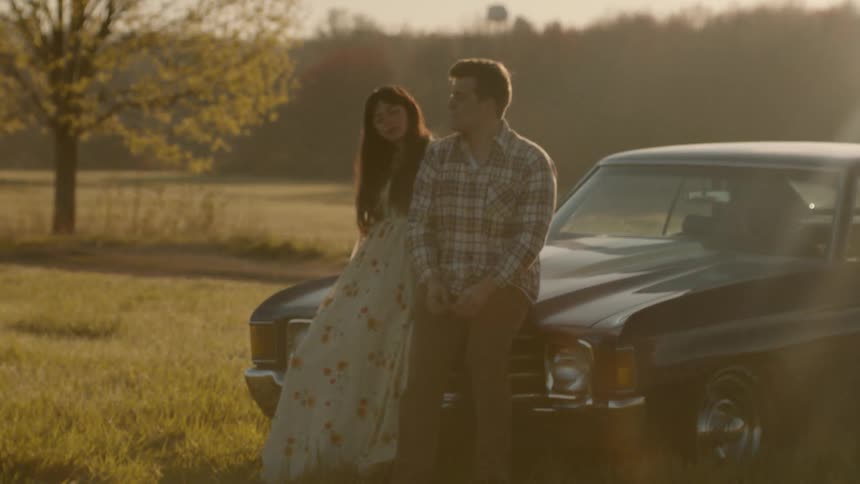Man and woman sitting against hood of car while sun sets