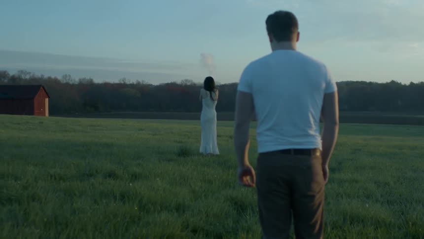 A man watches a woman standing in the grass with her back to him