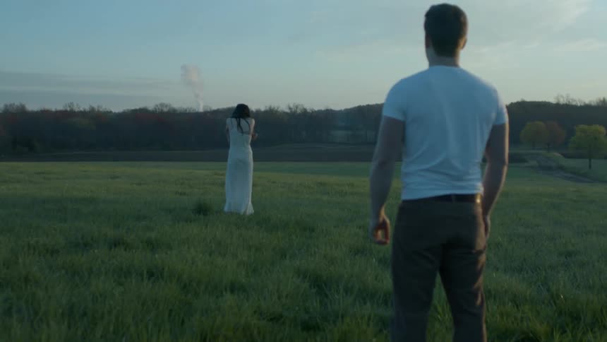 A lady in a white gown is looking in the distance as a man in a white shirt stands behind her