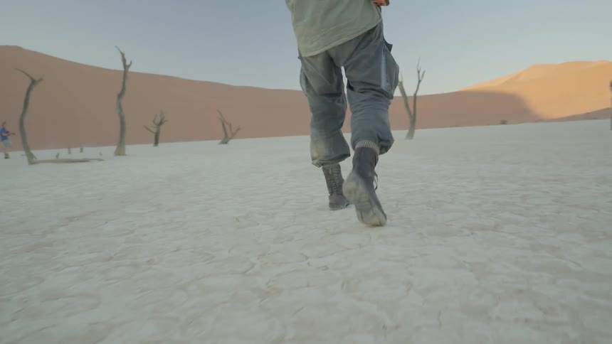 A man with a camera is walking in a desert landscape dotted with dead trees