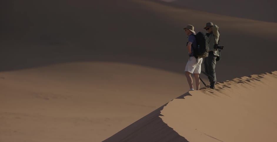 Two men carrying cameras and packs stand on top of a sand dune