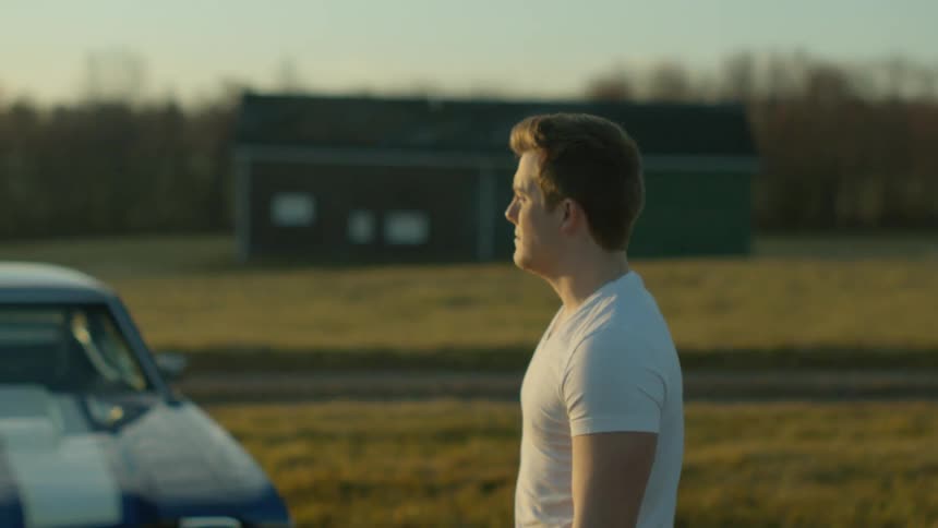 Man standing outdoors in a t shirt next to his muscle car in the country