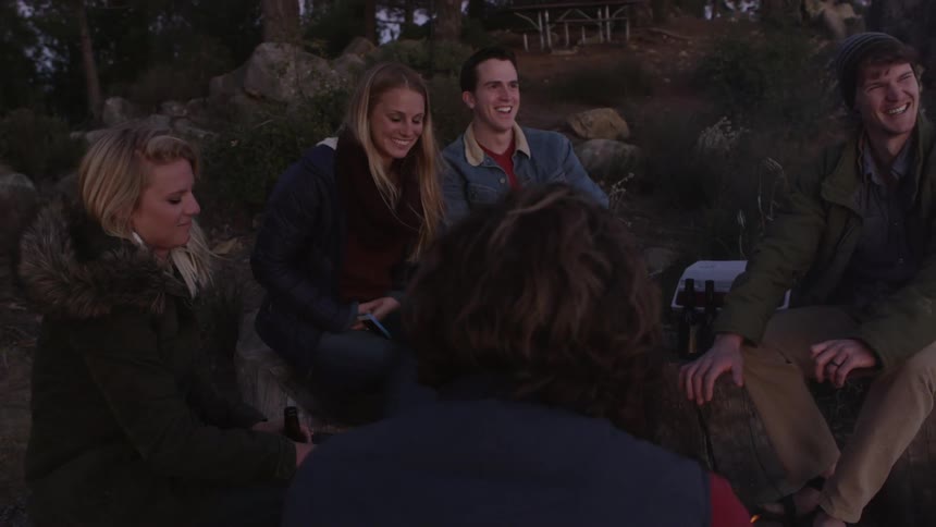 A group of friends sit around a campfire laughing and talking