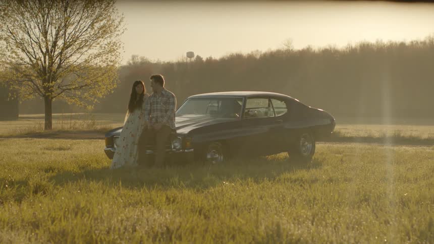 A man and woman are sitting on the hood of a car in the middle of a field