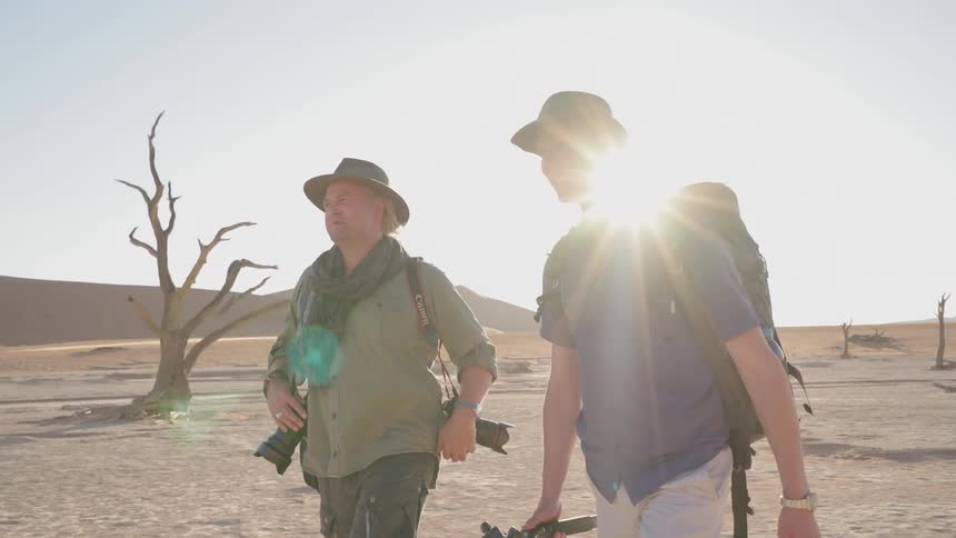 Two photographers carrying equipment walk in the desert