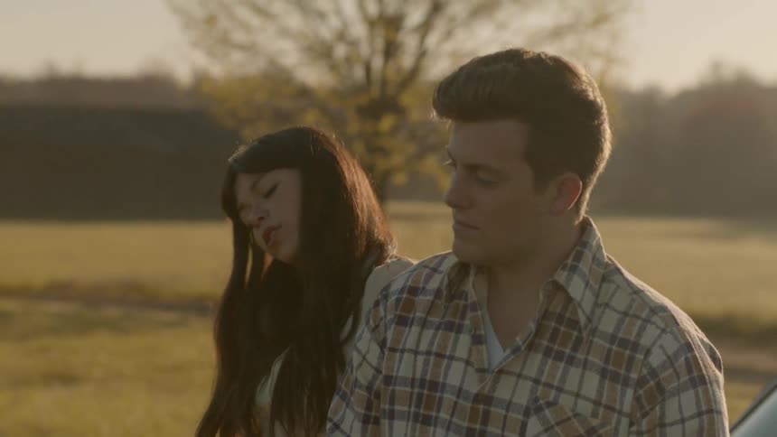 A man and woman a talking as they sit on a car outside at dawn