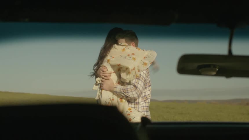 A man and woman embrace in an open field in front of a car