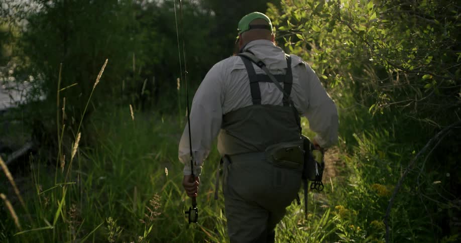 A man in waders carries a fishing pole as he walks a grassy trail