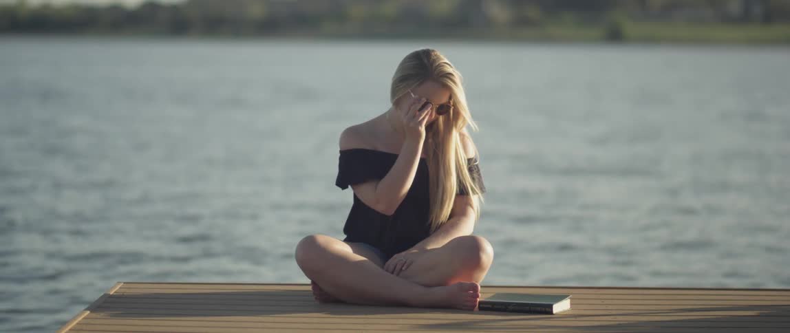 A blonde haired woman sits on a lake dock with a book