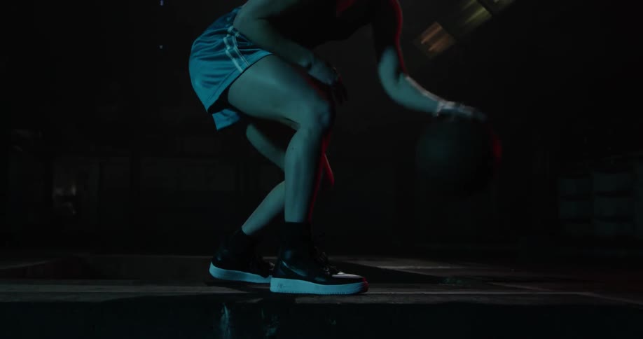 A woman is practice dribbling her basketball in a dark alley