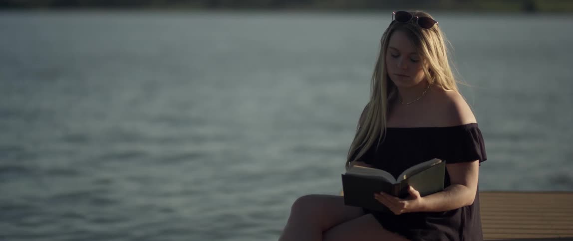 A woman sitting near the ocean pauses from reading a book
