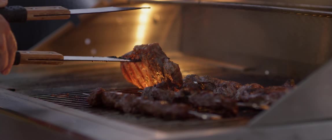 A person uses tongs to turn and check the meat on a grill