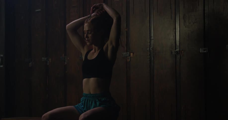 A athlete in a locker room removes her shirt and ties up her hair