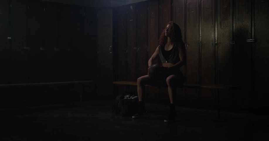 A woman plays with a basketball as she sits in a locker room