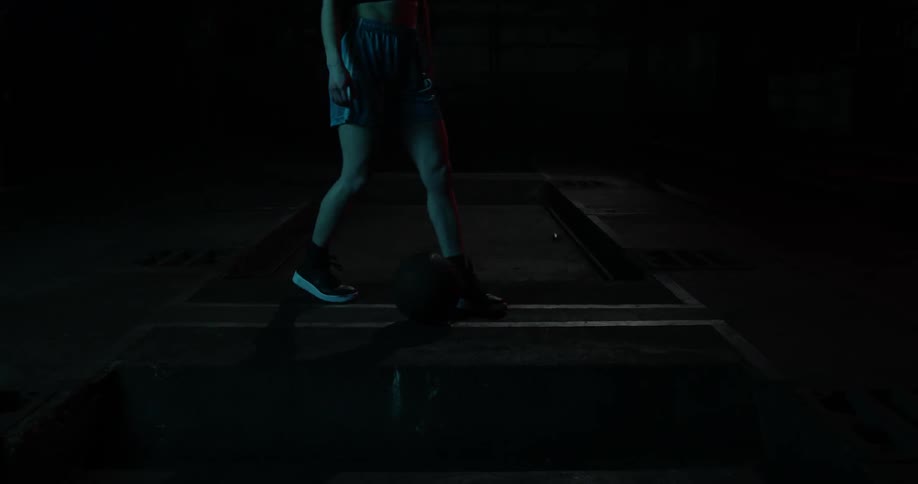 A woman crouches and lifts a basketball in the dark