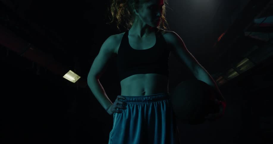 A woman stands with a basketball on her hip in a dark room
