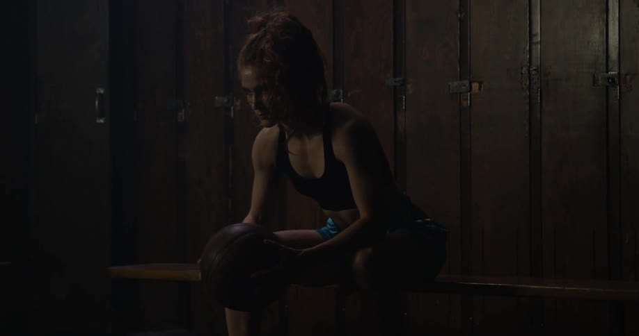 A woman sits on a bench gripping a basketball and stands up
