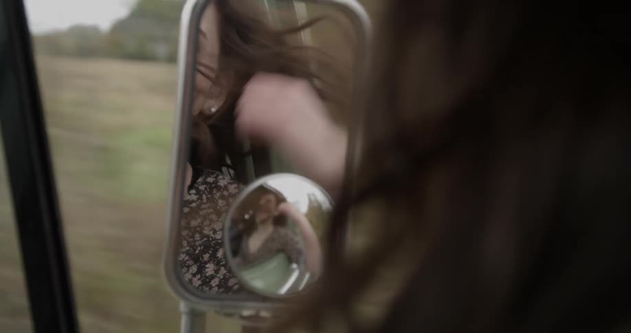 A woman's face is reflected in a side view mirror while traveling in the countryside