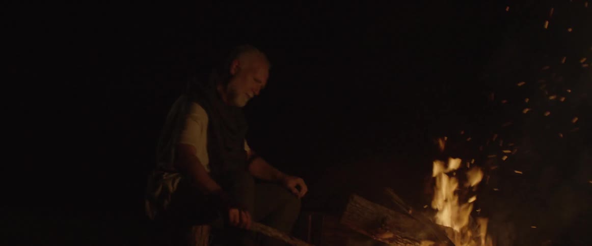 A man tends to the camp fire with a stick then adds a log