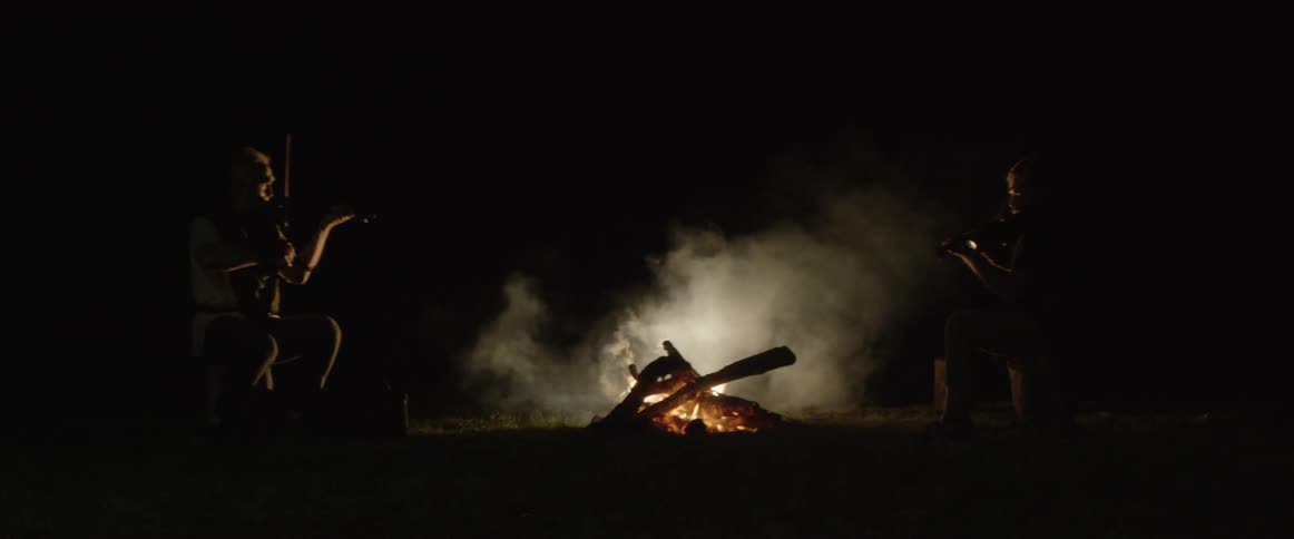 Two men play violin in front of a fire
