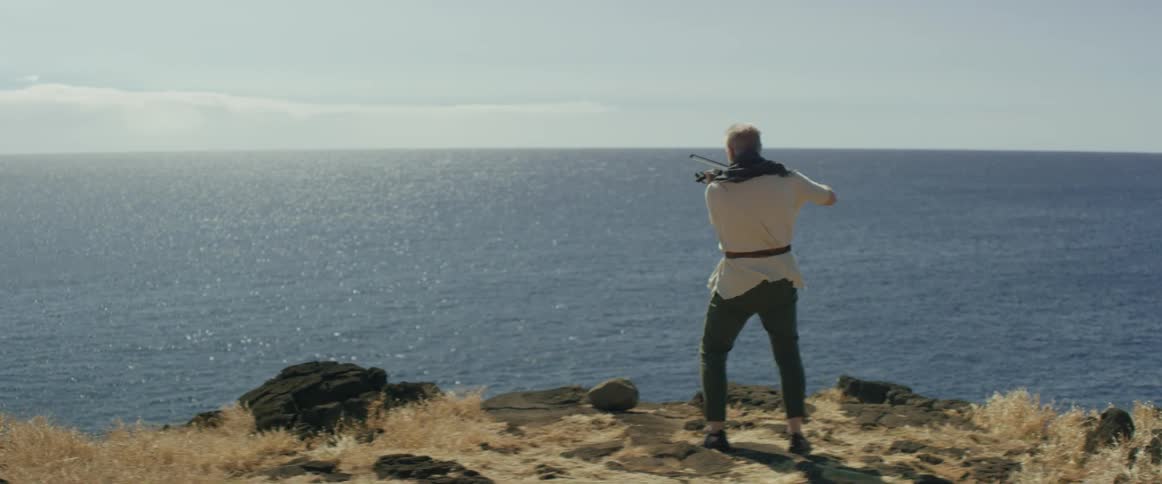 A man in a white tunic is playing the violin by the water