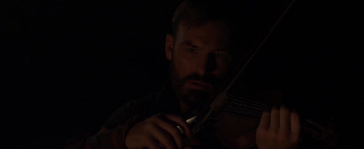 The man looks around in the dark by the campfire before playing violin