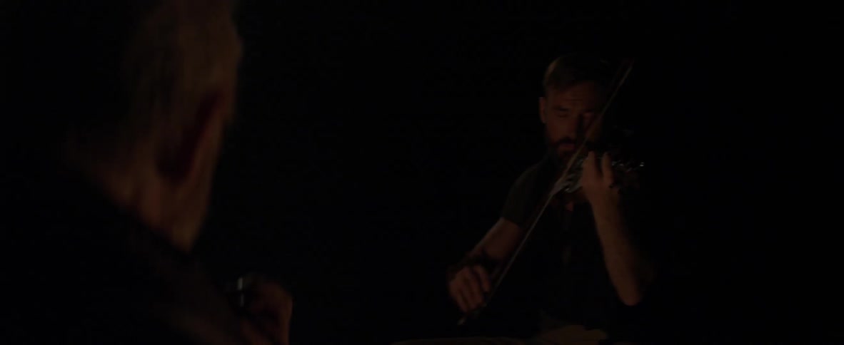 Two men play violin next to a campfire