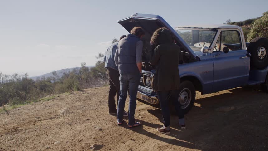 Two men and a woman contemplate the engine of a broke down late model pickup truck by the side of the road