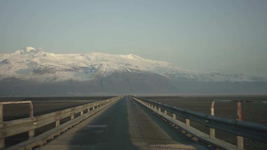 First person view of driving over a long bridge with snow capped mountains in the distance