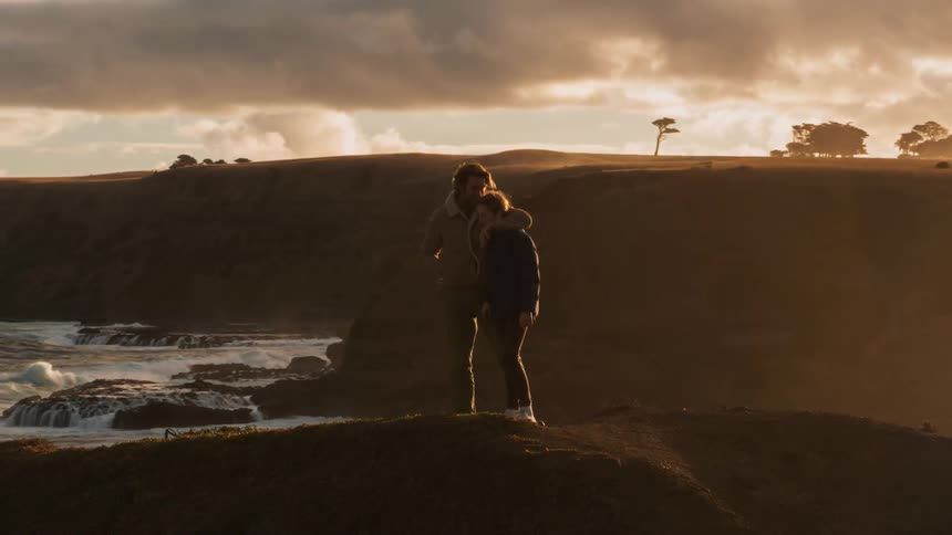 A young girl and a man are standing on the edge of a cliff next to the ocean