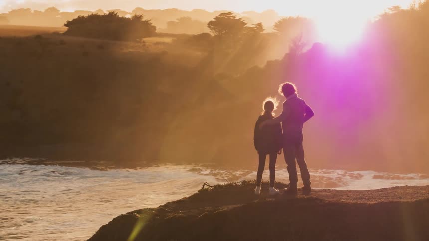 A man and girl stand on a cliff overlooking the ocean at sunset
