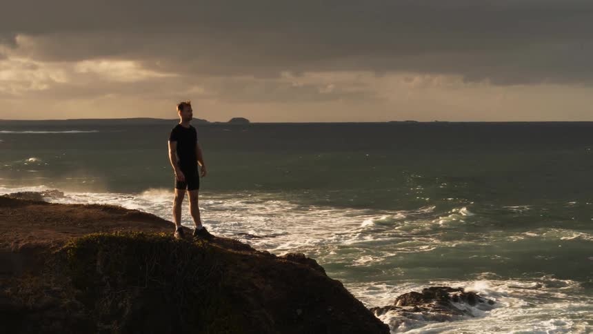 A man stands on the edge of a cliff by the water