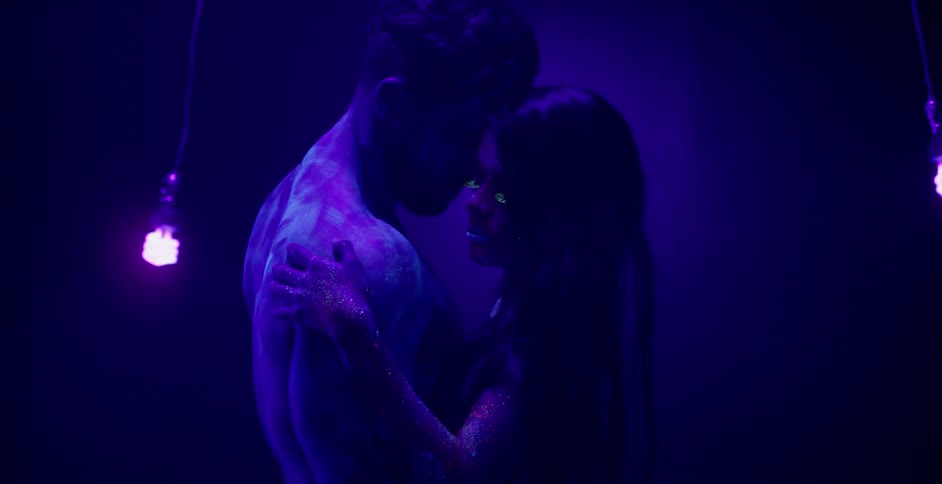A man and woman embrace in a black lit room